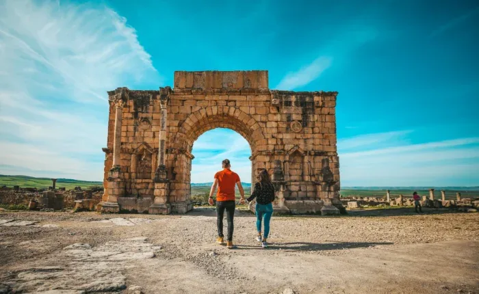 Two visitors walking toward an ancient stone arch under a blue sky
